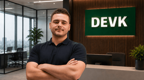 ein junger mann mit verschränkten Armen in einem dunklen Poloshirt in einem modernen Büro mit DEVK Logo