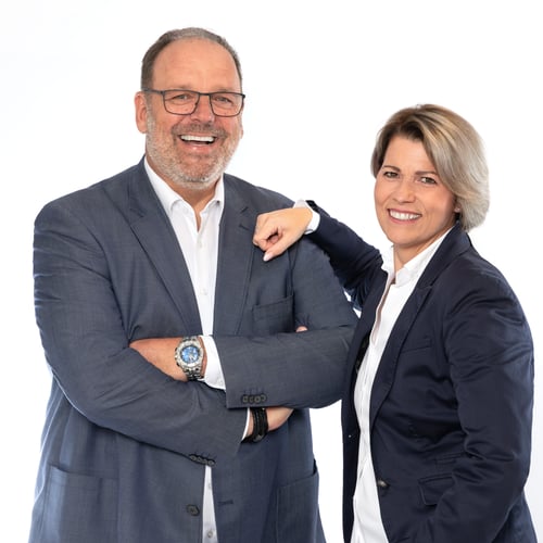 Das Foto zeigt links den Agenturleiter Marco Rütten stehend und rechts daneben steht seine Frau Heike Rütten, Mitarbeiterin in der Agentur. Sie steht ebenfalls und hat ihre rechte Hand auf seine linke Schulter gelegt. Beide lächeln.