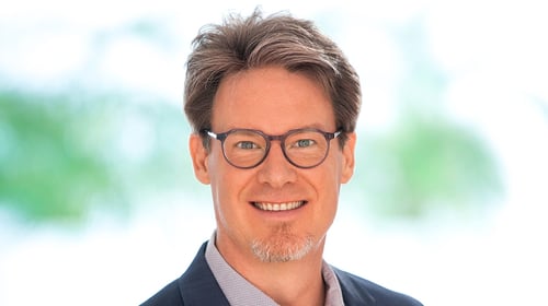 Porträt eines lächelnden Mannes mit kurzen braunen Haaren und rundem Brillengestell. Er trägt ein Hemd und ein dunkles Sakko. Im Hintergrund ist eine helle, weich unscharfe Umgebung mit grünen Farbtönen zu sehen.
