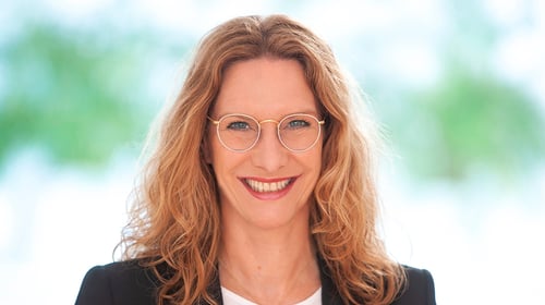 Porträt einer lächelnden Frau mit mittellangen, gewellten Haaren und runder Brille vor einem hellen, unscharfen Hintergrund.