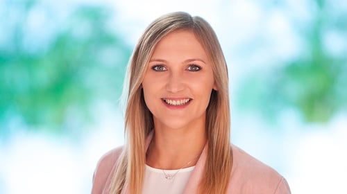 Nahaufnahme einer freundlich lächelnden Frau mit langen, glatten blonden Haaren. Sie trägt ein weißes Oberteil, eine zartrosa Jacke und eine dezente Halskette.