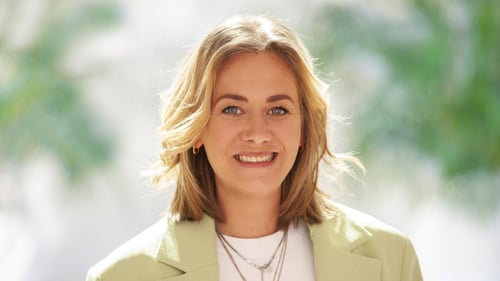 Porträt einer lächelnden Person mit schulterlangem blondem Haar, die direkt in die Kamera blickt. Sie trägt ein helles Oberteil und ein hellgrünes Sakko. Der Hintergrund ist weich unscharf mit grünen und hellen Farbtönen.