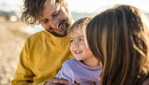 Nahaufnahme einer kleinen Familie am Wasser. Ein Kind lehnt den Kopf an die Schulter einer erwachsenen Person.