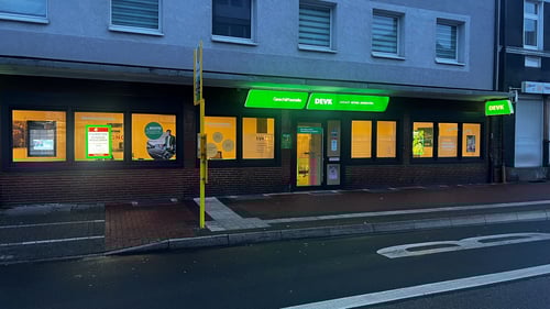 Das Foto zeigt die DEVK Geschäftsstelle Leandros Mpasouris in Recklinghausen Süd von Außen. Es ist früh am Abend und man sieht leuchtende Werbung.