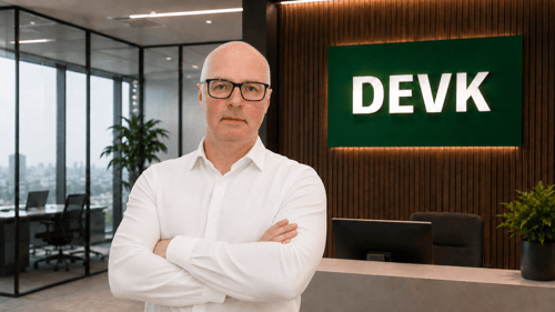 Das Bild zeigt einen Mann mit Brille in einem weißen Hemd. Im Hintergrund sieht man ein Büro mit DEVK logo