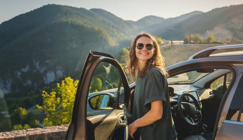 Frau mit Sonnenbrille steigt glücklich aus dem Auto aus und im Hintergrund erstreckt sich eine Berg- und Tallandschaft.
