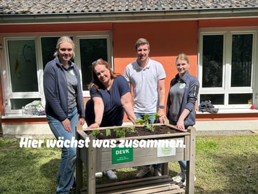Vier Personen stehen lächelnd um ein bepflanztes Hochbeet mit DEVK- und „Junges Gemüse"-Schild im Außenbereich des Johanniter-Kindergartens Grötzenberg. Im Beet wachsen erste Setzlinge wie Erdbeeren und Petersilie. Im Vordergrund der Schriftzug: „Hier wächst was zusammen."