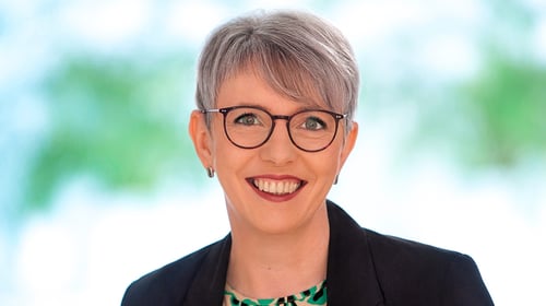 Nahaufnahme einer freundlich lächelnden Frau mit grauem Kurzhaarschnitt und dunkler Brille. Sie trägt einen schwarzen Blazer über einer grün-schwarz gemusterten Bluse.