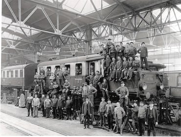 Zu sehen ist ein schwarz weiss Foto mit einem alten Triebwagen. Auf und vor dem Triebwagen sitzen und stehen Arbeiter.  
Das Foto wurde in einem Bahnhof aufgenommen.