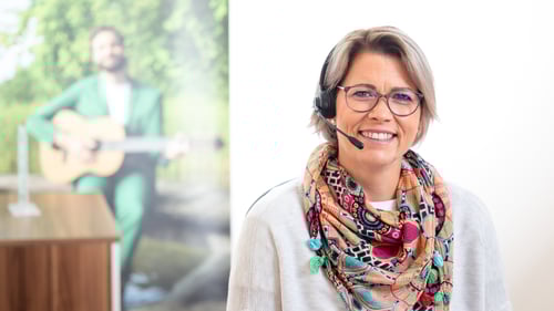 Das Bild zeigt Heike Rütten mit Headset und einem Lächeln an ihrem Schreibtisch