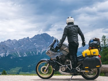 Im Vordergrund ist ein Motorrad zu sehen auf dem der Fahrer steht und in die Berge im Hintergrund schaut. Das Motorrad ist mit Gepäck beladen und vermittelt den Eindruck einer Tour.