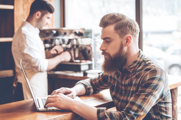 Ein Mann mit Hemd und Bart sitzt mit einem Laptop an einer Theke eines Cafés. Im Hintergrund steht ein Barista, der eine Kaffeemaschine bedient.