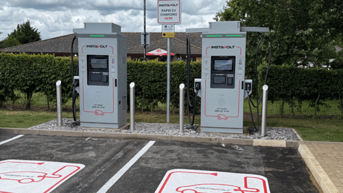 Two InstaVolt rapid EV chargers in a dedicated charging bay with clear ground markings, set in a green roadside location with hedges and trees.