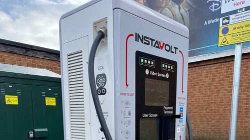 InstaVolt rapid EV charging station at a service station, featuring CCS connectors, contactless payment terminal and user screen, with signage and brick wall in the background.