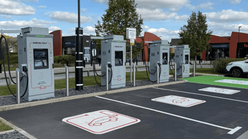 Row of InstaVolt ultra-rapid EV chargers in a modern car park with clearly marked electric vehicle bays, surrounded by shops and greenery