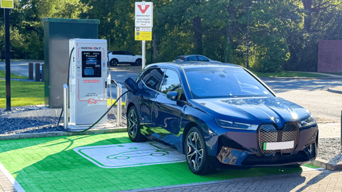 A BMW electric vehicle charging at an InstaVolt ultra-rapid charger in a green-marked EV bay beside a quiet road and wooded area.