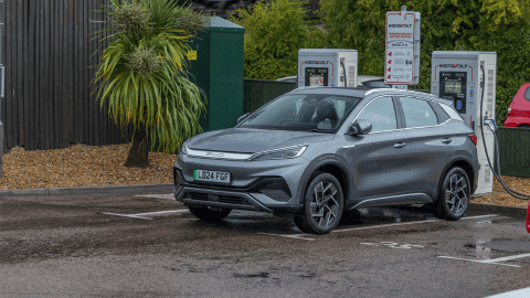 A sleek grey electric car charging at an InstaVolt station, set in a wet car park with tropical-style plants and a moving train visible in the background.