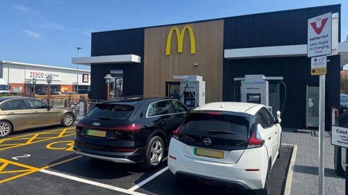 A pair of InstaVolt ultra‑rapid electric vehicle (EV) charging units installed side by side in a marked parking bay at a roadside location. Each charging unit is sleek, rectangular, and light‑coloured with a display panel and coiled charging cables attached. The bays are clearly delineated with painted symbols and lines on the tarmac. The overall scene reflects a clean, organised installation typical of an EV charging site at a McDonald’s or retail park near major roads such as the A295.