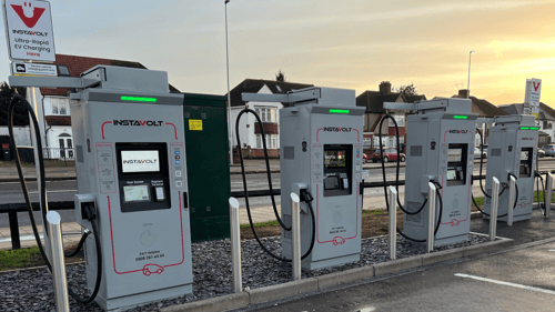 InstaVolt ultra-rapid EV chargers installed in a roadside car park, showing charging screens, cables and protective bollards, with a suburban street behind.