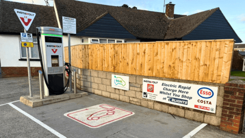 InstaVolt rapid charging station with adjacent signage for amenities, including Esso, Costa, Spar, and MG. Marked EV parking bay and wooden fencing in a well-maintained parking area.
