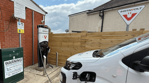 A white electric van charging at an InstaVolt rapid EV charger next to a brick building, with clear signage and CCTV warning notices visible.
