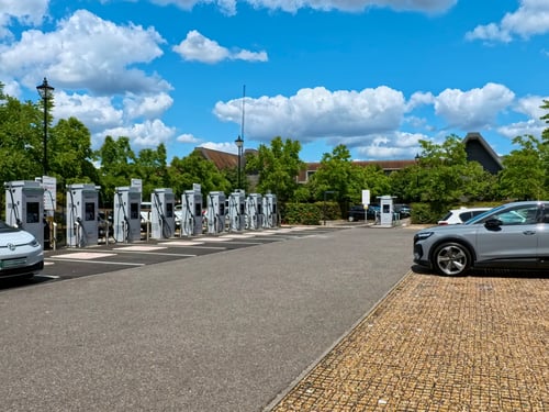 Array of InstaVolt rapid electric vehicle charging stations at Syon Park car park in London under a clear blue sky with fluffy clouds, featuring multiple chargers and parked electric cars, showcasing accessibility and modern infrastructure for eco-friendly transportation.