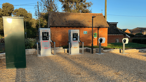 InstaVolt ultra rapid EV chargers and AC charging units at Beach House, Park Lane, Lymington. A large green utility cabinet is visible on the left, and a red-brick building with a sloped roof and a defibrillator box is in the background.