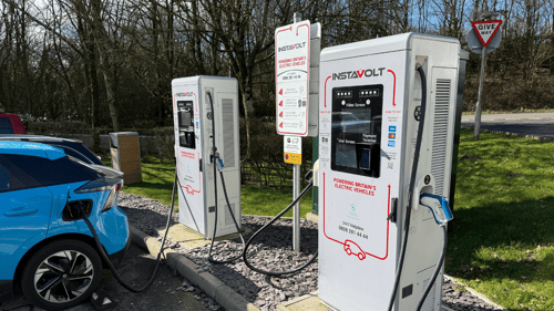 Two InstaVolt ultra rapid EV chargers in a parking area, with a blue electric vehicle plugged in. A give way sign is visible in the background along with trees and grass.