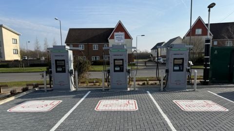 A wider view of three InstaVolt EV charging stations in a paved parking area with designated charging spots marked with red and white painted symbols. The surrounding environment includes residential houses, streetlights, and a road. A sign above the chargers indicates "InstaVolt Rapid EV Charging Here.