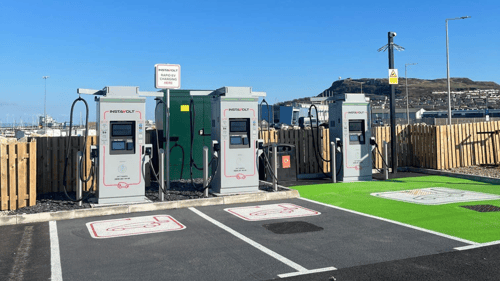 InstaVolt ultra rapid EV chargers at Portland - Starbucks, located on Hamm Breach Road, Portland, DT5 1DX. The charging bays are marked with green-painted areas, and the backdrop features a scenic coastal view with hills and a marina.