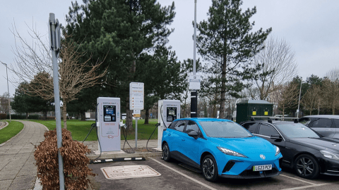 Two InstaVolt ultra rapid EV chargers installed in a well-maintained car park, with a blue electric car actively charging. The site is surrounded by trees and a pedestrian pathway nearby.