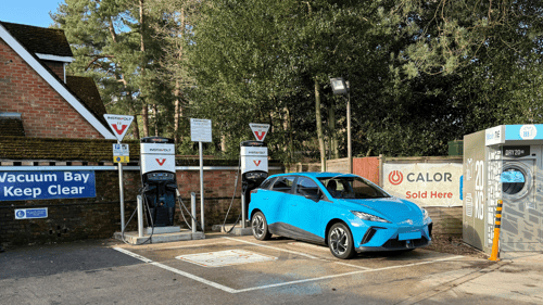 A bright blue electric car charging at InstaVolt EV chargers at Cresswells Garage, Wokingham, surrounded by trees and signage.