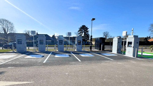A wide-angle view of an InstaVolt charging site featuring six ultra rapid EV chargers, marked parking spaces, and a black equipment unit against a suburban backdrop.