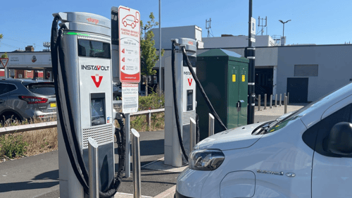 Two InstaVolt rapid EV chargers installed in a retail car park, with a white electric van plugged in and charging. Signage details contact information and charging instructions. Nearby cars and buildings are visible under a clear blue sky.