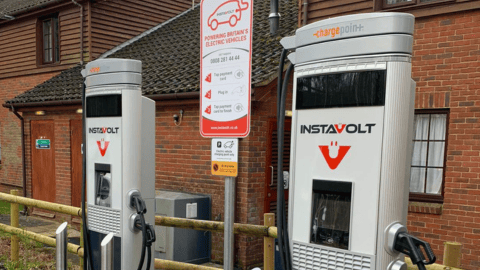 Two InstaVolt ultra rapid EV charging units situated outdoors next to a brick building, with a sign displaying charging instructions and payment options. A wooden fence and a small landscaped area are visible in the foreground.