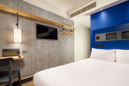 landscape image of modern bedroom with double bed in foreground covered in smooth white linen and four plump white pillows against a blue headboard. to the side of the bed is a small desk with chair and flat screen television, in the background is a coat rail with hangers. The walls are grey wood panelling.