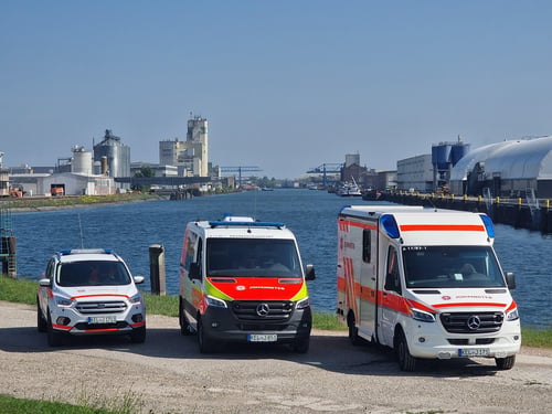 Drei Einsatzfahrzeuge der Johanniter in Kehl vor der Kulisse des Kehler Rheinhafens. Rechts ein Rettungswagen, in der Mitte ein Krankenwagen und links ein PKW mit Blaulicht.